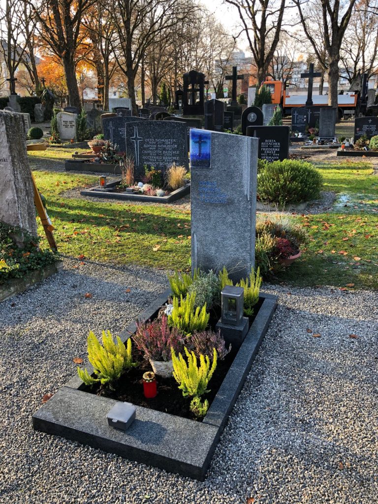Grabstein aus grauem Granit mit rechteckiger blauer Glaseinlage und Kreuzornament in herbstlichem Friedhofsambiente