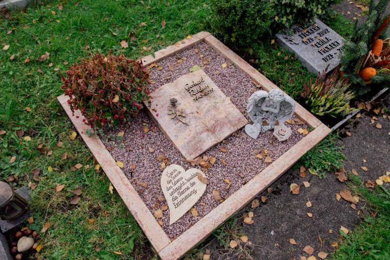 Urnengrab-Liegestein in rosa Naturstein mit Buch-Motiv, Engel-Figur und herzförmigem Stein auf Kiesanlage